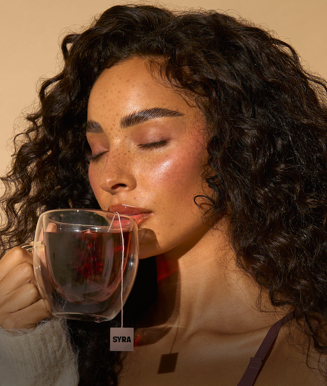 Woman drinking from a clear mug with a visible 'SYRA' brand label against a beige background